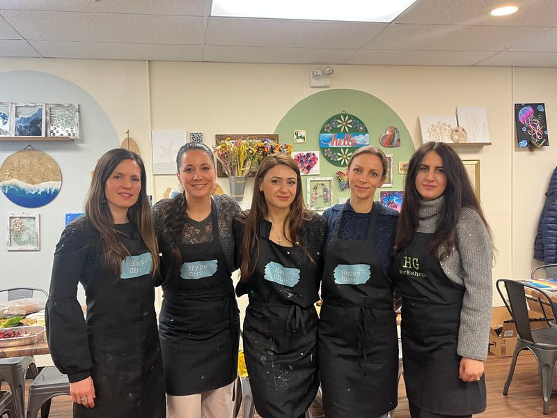 Five women in black aprons standing together in a room with art supplies and decorations on the walls.